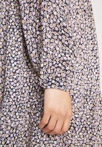 Main de femme reposant à côté de la manche d'une robe en tissu floral foncé présentant de petites fleurs roses, blanches et jaunes sur fond bleu marine.