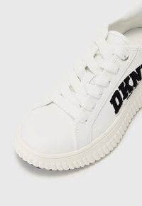 Sneaker bianco con una parte superiore sintetica liscia, punta rotonda e suola in gomma texturizzata. Logo "DKNY" nero sul lato. Lacci piatti.