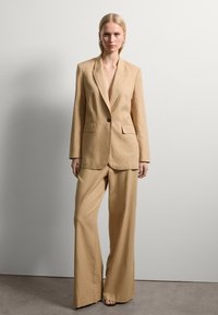 Beige Leinenanzug mit einem Ein-Knopf-Blazer und weitem Hosenbein. Besitzt einen tiefen Ausschnitt und zwei Vordertaschen am Blazer.
