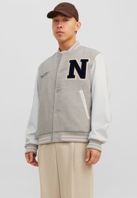 Grijze en witte varsityjacket met een geborduurde marineblauwe "N," drukknoopjes, ribbelrand en contrasterende leren mouwen.