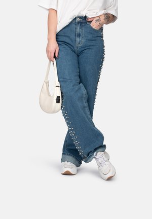 Jeans van denim met een hoge taille, wijde pijpen en parelversieringen langs de zijkanten. Gecombineerd met een witte tas en sneakers.