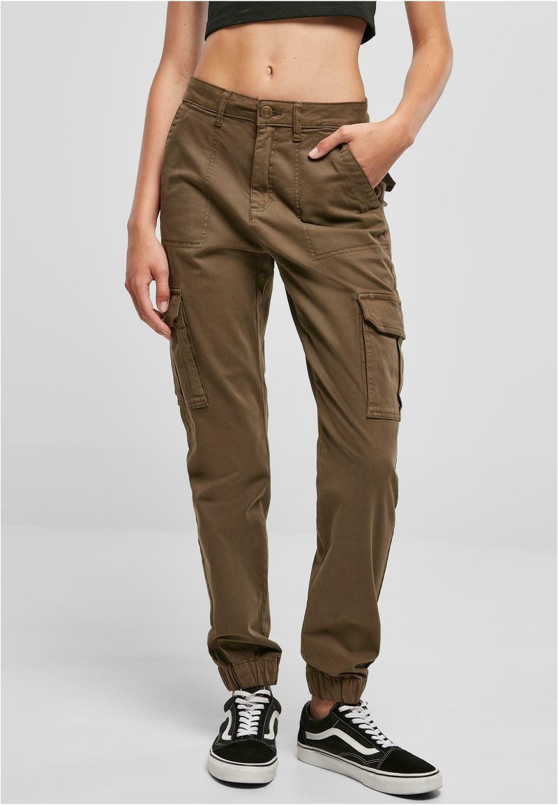 Woman wearing brown cargo pants with front pockets and elastic cuffs, paired with black and white sneakers, standing against a plain background.