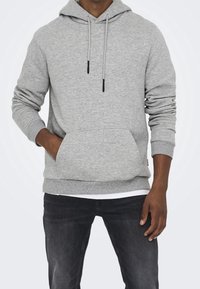 Szara bluza z kapturem wykonana z miękkiego materiału, z przednią kieszenią typu kangurka, regulowanymi troczkami, ściągaczami na mankietach i u dołu, noszona na ciemnych dżinsach.
