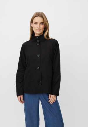 Frau mit glattem, hellbraunem Haar, die eine schwarze, geknöpfte Jacke mit hohem Kragen und lockere blaue Hosen vor weißem Hintergrund trägt.