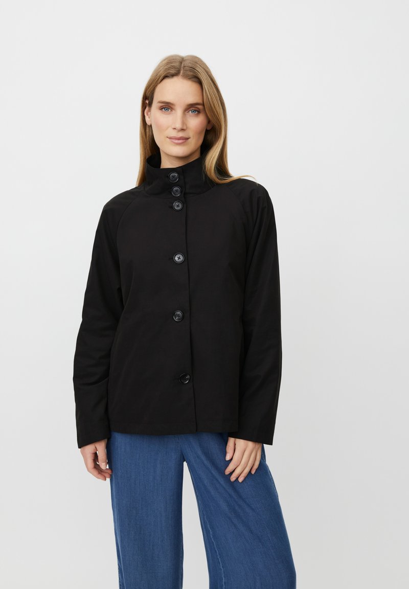 Žena s rovnými světle hnědými vlasy, která má na sobě černou zapnutou bundu s vysokým límcem a volné modré kalhoty, na bílém pozadí.