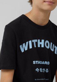 Schwarzes Baumwoll-T-Shirt mit fettem blauen Text, der "OHNE" sagt, und kleinerem Text darunter; runder Ausschnitt, kurze Ärmel.