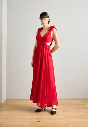 Red sleeveless dress with ruffled shoulder accents, a fitted waist, flowing skirt, and cut-out detail. Worn with black pointed-toe heels.