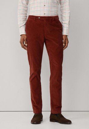 Cords in einem tiefen Rot, mit geradem Schnitt, seitlichen Taschen und einem Knopfverschluss. Getragen mit einem karierten Hemd.