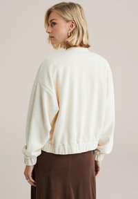 Krémový sveter s textúrou, voľného strihu, s rebrovaným lemom a nafúknutými rukávmi, v kombinácii s hnedou sukňou. Zobrazený zozadu.