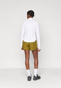 Λευκή μακρυμάνικη μπλούζα, ελιά πράσινα σορτς, μαύρα αθλητικά παπούτσια και λευκές κάλτσες. Διαθέτει ψηλό γιακά και αθλητική εφαρμογή.