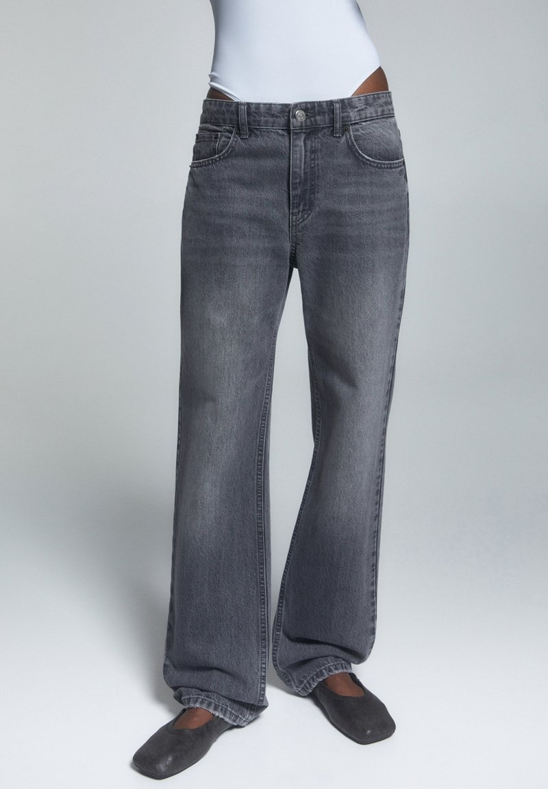 Personne portant un jean gris foncé ample, un haut blanc et des chaussures plates noires sur un fond clair uni.
