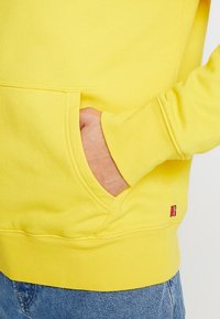 Sudadera amarilla con un bolsillo tipo canguro y puños acanalados. Presenta una tela texturizada y una pequeña etiqueta de marca roja en el dobladillo.