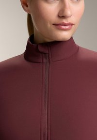 Tamnocrvena majica s visokim ovratnikom, izrađena od glatkog materijala, središnjim patentnim zatvaračem i bezšavnim dizajnom za elegantan izgled.