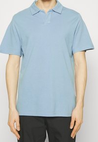 Muž ve světle modrém jednobarevném polo tričku s krátkým rukávem a černých kalhotách, stojící na neutrálním pozadí.