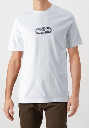 Bílý krátký rukáv tričko s logem "HILFIGER NEW YORK CITY 1985" v černé, červené a modré barvě, nošené s tmavě hnědými kalhotami.
