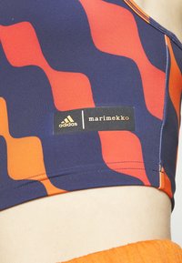 Primo piano di un top sportivo con un motivo ondulato arancione e blu scuro, che presenta i loghi di Adidas e Marimekko sul tessuto.