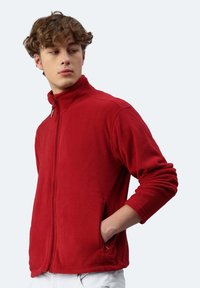 Giacca in pile rossa con colletto alto, zip completa e tasche laterali. Tessuto morbido e vestibilità rilassata con maniche lunghe.