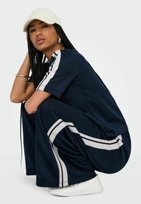 Marineblaues Athletik-Set mit einem kurzärmlichen Oberteil und weiten Hosen mit weißen Seitstreifen. Texturiertes Gewebe und weiße Mütze. Weiße Sneakers.
