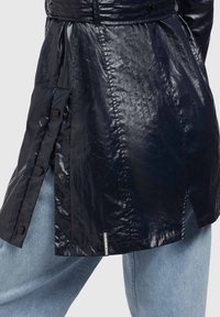 Námořnická modrá, lesklá trench coat s knoflíkovými bočními kapsami, páskem v oblasti pasu a texturovaným povrchem. Obsahuje malou logo nášivku na lemu.