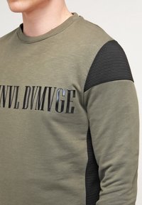 Olijfgroene sweatshirt met geribbelde zwarte schouderaccenten en zwarte bedrukte tekst. Heeft een ronde hals en een aansluitend ontwerp.