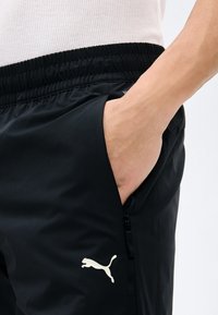 Χέρι στην τσέπη μαύρου παντελονιού φόρμας Puma με ελαστική μέση και τσέπη με φερμουάρ, φορεμένο με λευκό ριμπ τοπ.