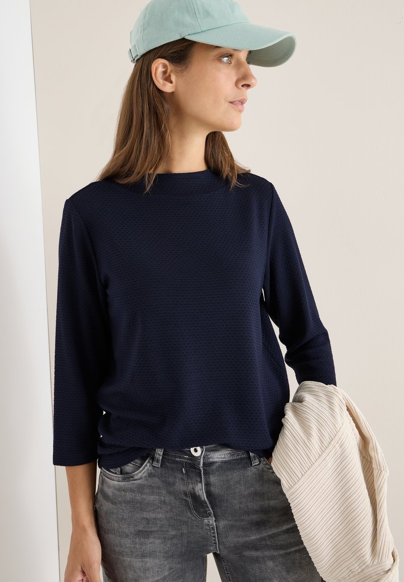 Marineblauw textuur long-sleeve top met hoge hals, gecombineerd met grijze jeans. Een lichtgekleurde gestreepte item wordt in één hand vastgehouden.