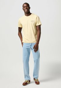 Camiseta de algodón amarilla con mangas cortas, que presenta un pequeño logo en el pecho, combinada con jeans de color azul claro y zapatillas marrones con acentos amarillos.