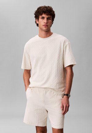 Mann mit lockigem Haar, der ein cremefarbenes, gesteppte Kurzarmhemd und passende Shorts trägt, steht mit einer Hand in der Tasche vor grauem Hintergrund.