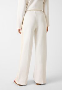 Pantaloni a gamba larga in tessuto crema con un pannello laterale testurizzato. Presentano una vita regolabile con cordino e una texture liscia e morbida. Accenti rossi all'orlo.