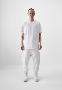 Fehér, túlnyúlt póló kerek nyakkivágással, fehér, oldalzsebes, lazán illeszkedő jogger nadrággal párosítva, fehér sportcipővel viselve.