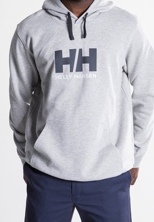 Mann trägt hellgrauen Helly Hansen Hoodie mit großem HH-Logo und Schriftzug, dazu dunkelblaue Hose, Hände in den Taschen.
