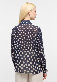 Marineblaue transparente Bluse mit großen cremefarbenen Punkten, langen Ärmeln und Kragen. Verfügt über einen abgerundeten Saum und Knopfmanschetten.