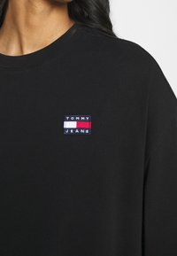 Svart bomullströja med rund halsringning, med en liten rektangulär broderad logotyp i blått, vitt och rött på vänster bröst.