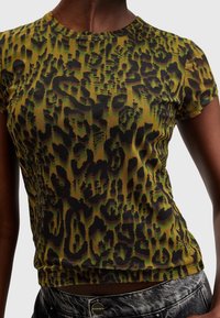 Närmast av en person som bär en åtsittande grön och svart leopardmönstrad kortärmad topp tillsammans med blekta svarta jeans.