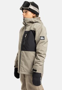 Osoba ubrana w beżową i czarną zimową kurtkę, czarną wełnianą czapkę z goglami narciarskimi i czarne rękawiczki, zwrócona w lewo na białym tle.