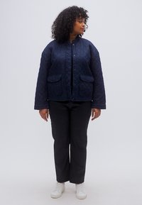 Ναυτικό μπουφάν καπιτονέ με στρογγυλή λαιμόκοψη, κούμπωμα με κουμπιά τύπου snap και δύο εμπρόσθιες τσέπες, σετ με μαύρο παντελόνι και λευκά αθλητικά παπούτσια.