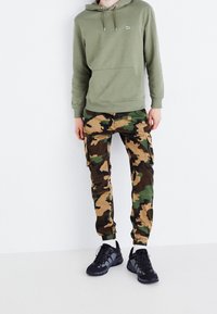 Persoon in een olijfgroen hoodie en camouflage cargo-broek met zwarte sneakers, staande tegen een witte muur.