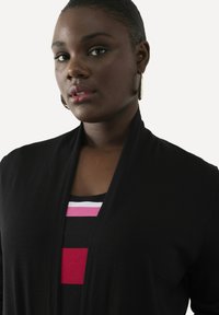 Schwarzer Cardigan mit einer weichen Textur, offener Front und langen Ärmeln, über einem gestreiften Hemd mit pinken, weißen und schwarzen Streifen getragen.