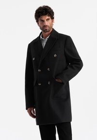 Abrigo negro de lana de corte cruzado con grandes botones, cuello con muescas, bolsillos laterales y un acabado ligeramente texturizado.
