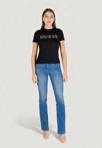 Camiseta negra ajustada con el logo "GUESS" en dorado metálico, combinada con vaqueros de pierna recta en azul claro y tacones altos en tono nude.