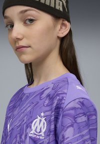 Maglietta sportiva viola con grafiche testurizzate, colletto rotondo a giro, e emblema bianco. Presenta un tessuto simile a rete sulle spalle.