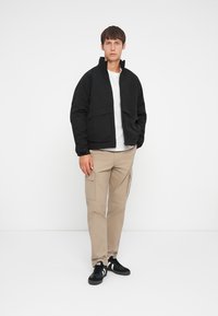 Black zip-up jacket with high collar, round hem, and two front pockets. Worn over a white shirt, paired with beige cargo pants and black sneakers.