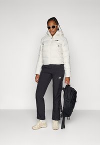 Hvit pufferjakke med glidelåslommer, svarte bukser med logo ved kneet, beige joggesko og en svart ryggsekk med justerbare stropper.