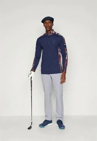 Cămașă de golf cu mâneci lungi bleumarin, cu mâneci cu model, guler cu fermoar, pantaloni gri deschis, încălțăminte bleumarin și o pălărie asortată, ținând un club de golf.