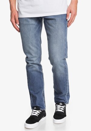 Jeans in denim azzurro chiaro con un design a gamba dritta, caratterizzati da una leggera sbiaditura, tasche laterali e bordi consumati, abbinati a sneakers nere.