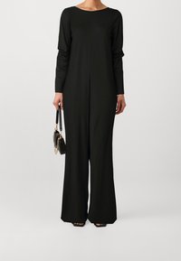 Mono negro de manga larga con escote barco y diseño de pierna ancha. El material parece suave; complementado con un pequeño bolso negro.