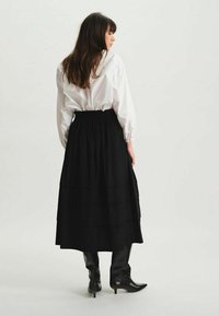 Jupe maxi noire à volants avec taille smockée, assortie à une chemise blanche à manches longues. Le modèle porte des bottes noires montantes.