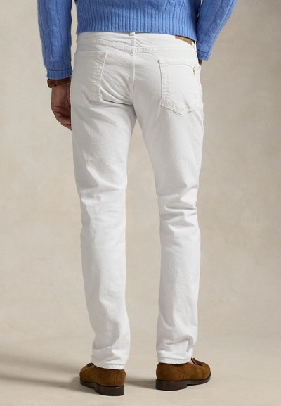 Muž nosící bílé džíny, hnědé semišové mokasíny a modrý svetr s copánkovým vzorem, stojící před neutrálním pozadím.