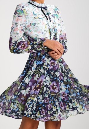Mujer que lleva un vestido floral de manga larga con múltiples estampados, con un cuello blanco con volantes y detalle de cinta negra.