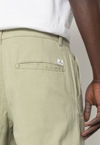 Mörkhyad person som bär ljusgröna byxor och en vit skjorta, visar bakfickan och sömmen på sidan med handen avslappnad vid sidan.